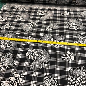 May include: Black and white plaid fabric with a repeating floral pattern. The flowers are white with a detailed, linear design. A yellow measuring tape is visible across the fabric. The fabric is rolled up in the background.
