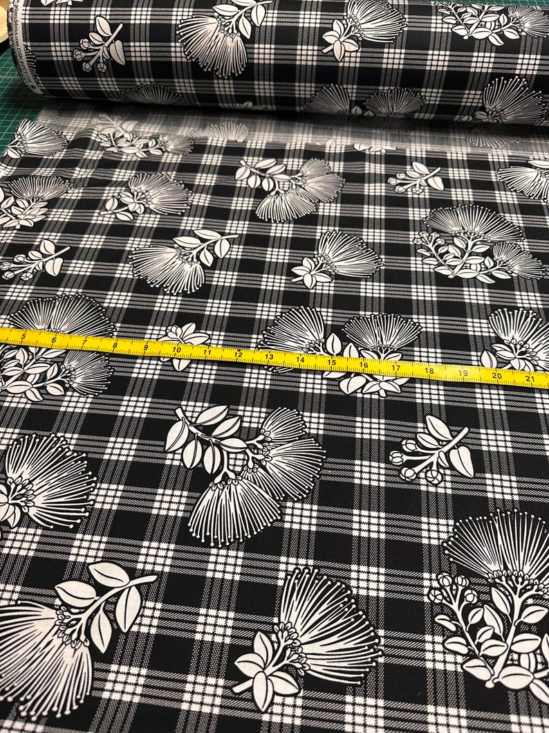 May include: Black and white plaid fabric with a repeating floral pattern. The flowers are white with a detailed, linear design. A yellow measuring tape is visible across the fabric. The fabric is rolled up in the background.