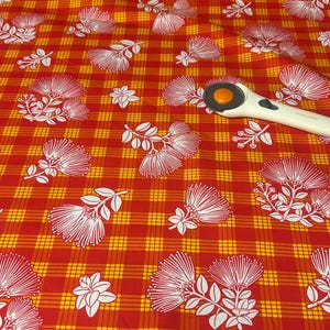 May include: A vibrant red and yellow plaid fabric featuring a repeating white floral pattern of pohutukawa blossoms. A white and orange rotary cutter is positioned on the fabric surface, providing a sense of scale for the printed textile design.