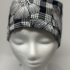 May include: A black and white patterned scrub cap. The cap features a plaid design with white floral accents. The cap is displayed on a mannequin head.