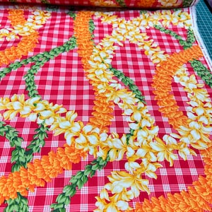 May include: Red and white checkered fabric with a repeating pattern of leis. The leis are composed of orange and yellow flowers, and green leaves. The fabric is rolled up in the background.