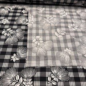 May include: Black and white plaid fabric with a floral design. The fabric features a repeating pattern of stylized white flowers and leaves against a black and white checkered background. The fabric is rolled up in the background.