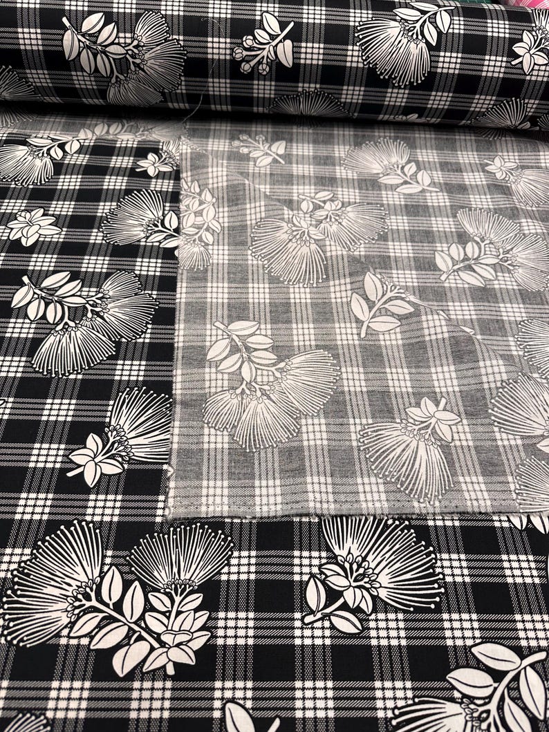 May include: Black and white plaid fabric with a floral design. The fabric features a repeating pattern of stylized white flowers and leaves against a black and white checkered background. The fabric is rolled up in the background.