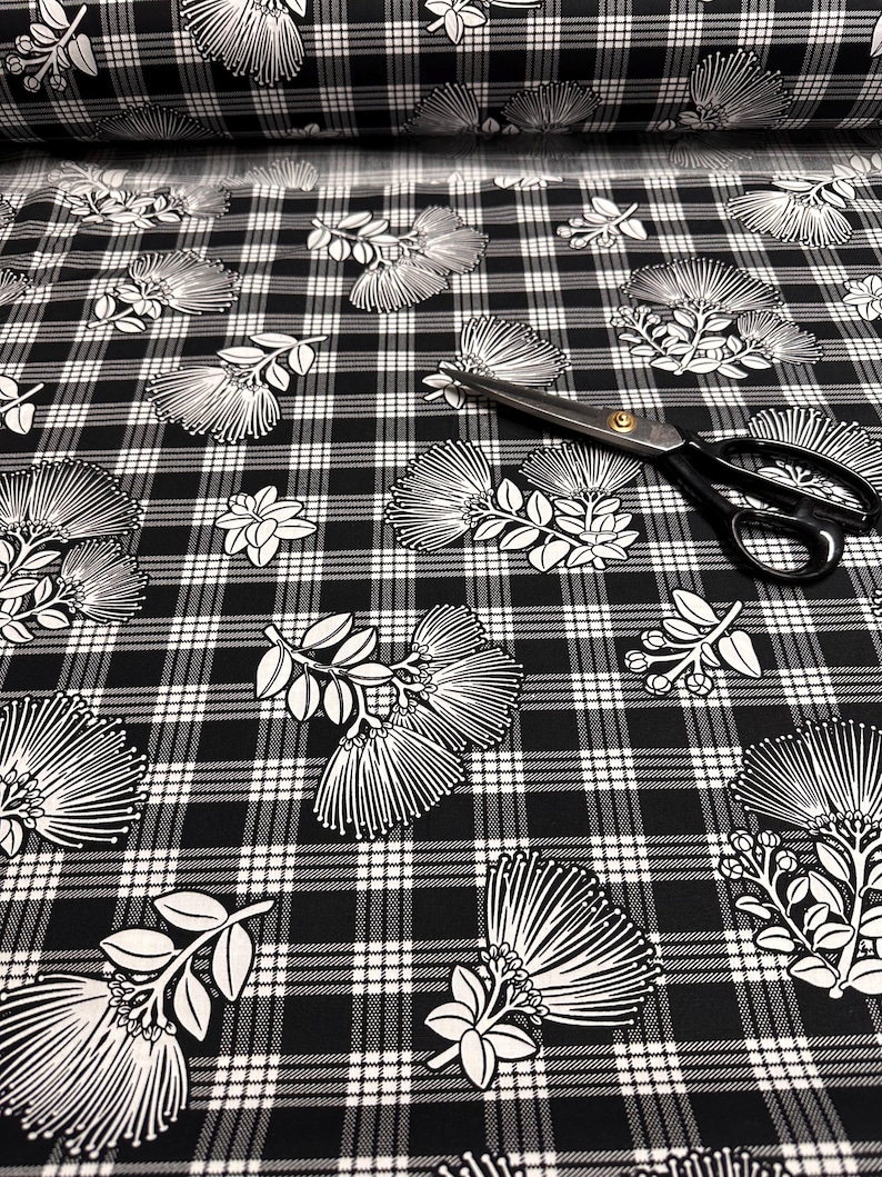 May include: Black and white plaid fabric with a floral design. The fabric features a repeating pattern of white flowers and leaves on a black and white checkered background. A pair of black scissors with gold accents rests on the fabric.