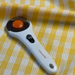 May include: A white rotary cutter with an orange blade and a black handle. The cutter is on a yellow and white checkered fabric. The handle has the word "FISKARS" printed on it.