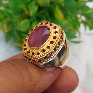 May include: A silver ring with a large oval-shaped pink gemstone surrounded by a gold setting with small red gemstones. The ring has a textured band.