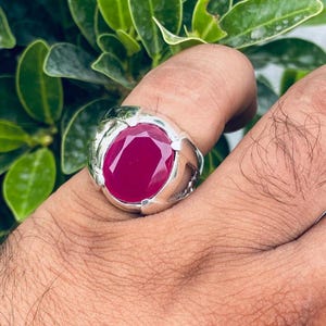 May include: A silver ring featuring a large, oval-cut, deep red gemstone. The ring has a detailed, textured design around the gemstone setting. The ring is worn on a finger, with green foliage in the background.