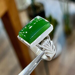 May include: A silver ring with a green gemstone set in a rectangular shape. The ring has a detailed design on the band.