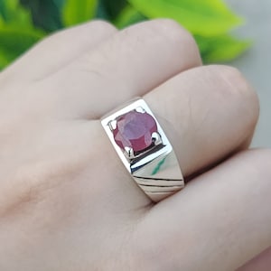 May include: A silver ring with a red gemstone set in a square bezel. The ring has a textured band.