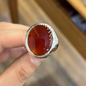Könnte beinhalten: Ein Silberring mit einem großen, ovalen Karneol-Edelstein. Der Stein hat eine tiefrot-orange Farbe und ist hochglanzpoliert. Das Ringband besteht aus hellem, poliertem Silber.