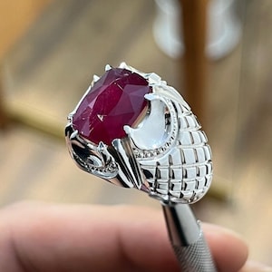 Peut inclure: Une bague en argent avec une grande pierre précieuse en rubis rouge sertie dans un design orné et détaillé.