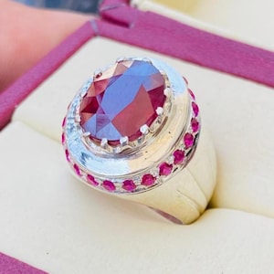 May include: A silver-toned ring featuring a large, oval-cut deep red gemstone at the center, surrounded by a halo of smaller, round-cut red stones set into the metal band. The ring has a wide, polished metallic shank and a substantial profile.