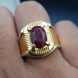 Könnte beinhalten: Ein gold-silberner Ring mit einem großen, oval geschliffenen Rubin. Der Rubin hat eine tiefrote Farbe und ist in einer goldenen Lünette gefasst. Der Ring hat ein goldenes Band mit strukturiertem Design.