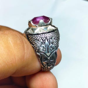 Op de afbeelding: Een zilveren ring met een grote, facetgeslepen, paarsrode edelsteen. De ring heeft een sierlijk, gedetailleerd ontwerp met bloemen- en textuurpatronen. De edelsteen is gezet in een kroonachtige zetting.