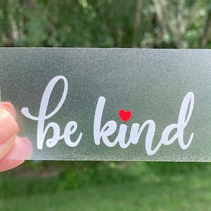May include: A frosted glass sticker with the text "be kind" in white cursive lettering and a small red heart above the word "kind".
