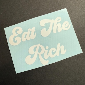 May include: A light blue sticker with the words "Eat The Rich" in a white, retro-style font. The sticker is rectangular and sits on a dark surface.