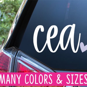 May include: White vinyl decal with the name "cea" in cursive script and a small light purple heart. The decal is on the back window of a car. The text "MANY COLORS & SIZES" is on a pink banner below the decal.