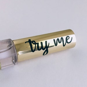 May include: A gold and clear tube with a white label that says "try me" in black cursive writing.