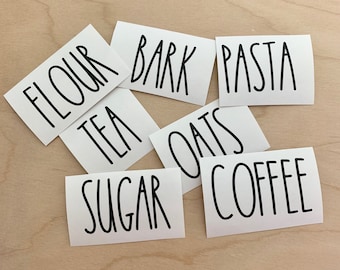 May include: White labels with black handwritten text for pantry organization. The labels read: "Flour", "Tea", "Sugar", "Bark", "Oats", "Pasta", and "Coffee".