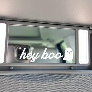 May include: A white ghost sticker with the text "hey boo" on a gray car visor.
