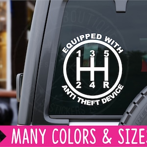 May include: White vinyl sticker with a black circle and the text "Equipped with Anti Theft Device". Inside the circle is a white gear shift pattern with the numbers 1, 2, 3, 4, 5 and the letter R.