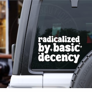 May include: White vinyl sticker applied to a vehicle window. The sticker displays the phrase "radicalized by basic decency" in a bold, stylized font with star accents. The sticker is designed for car windows and other smooth surfaces.