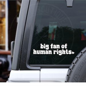 May include: White vinyl sticker with the text "big fan of human rights" in a bold, rounded font. The sticker is affixed to the rear window of a vehicle. The background is blurred, suggesting an outdoor setting.
