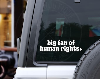 Può includere: Adesivo in vinile bianco con la scritta "big fan of human rights" in grassetto e arrotondata. L'adesivo è applicato sul lunotto posteriore di un veicolo. Lo sfondo è sfocato, suggerendo un ambiente esterno.