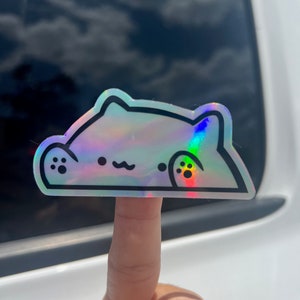 May include: A holographic sticker of a cartoon cat with a rainbow iridescent finish. The cat is in a curled up position with its paws visible.