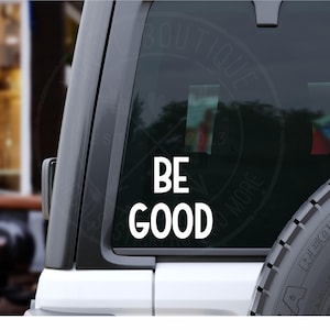 May include: White vinyl decal with the words "BE GOOD" in a bold, sans-serif font. The decal is applied to the rear window of a vehicle. The background is blurred, suggesting an outdoor setting.