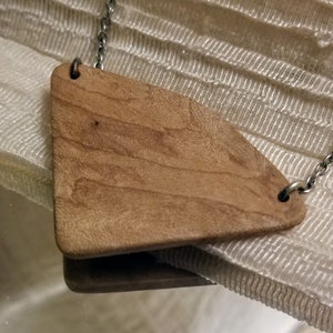 Puede incluir: Un colgante de madera en forma de diamante con un patrón de veta de madera natural cuelga de una cadena de plata. El colgante está unido a la cadena con dos anillos de plata.