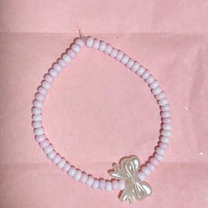 May include: A circular bracelet composed of small, round, light pink beads strung together. The bracelet features a single, translucent, off-white bow-shaped charm attached to the strand, creating a simple and uniform design throughout the piece.