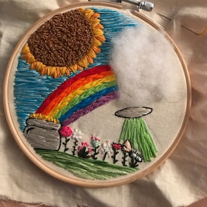 May include: Embroidered hoop art featuring a rainbow, a pot of gold, a sunflower, a cloud, a UFO, and flowers. The hoop is made of wood and has a white fabric background.