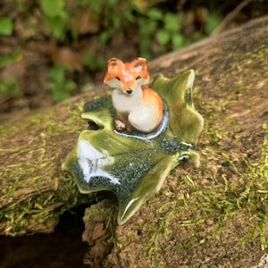 May include: A small ceramic figurine of a fox with orange and white fur, sitting on a glossy, dark green glazed ceramic leaf. The fox has pointed ears, a black nose, and a smooth, reflective finish, resting securely on the center of the leaf.