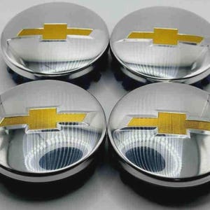 May include: Four chrome hubcaps with a gold Chevrolet bow tie logo. The hubcaps are round with a shiny, reflective surface and a black base. The gold logo is centered on each hubcap.
