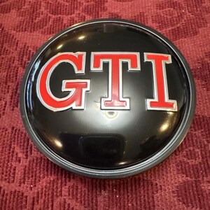 Puede incluir: Un emblema GTI negro y plateado con letras rojas. El emblema circular muestra las letras "GTI" en una fuente en negrita sin serifa con un contorno rojo. Acabado brillante, probablemente para un veh&iacute;culo.