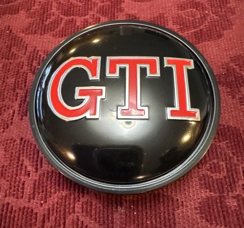 Puede incluir: Un emblema GTI negro y plateado con letras rojas. El emblema circular muestra las letras "GTI" en una fuente en negrita sin serifa con un contorno rojo. Acabado brillante, probablemente para un veh&iacute;culo.