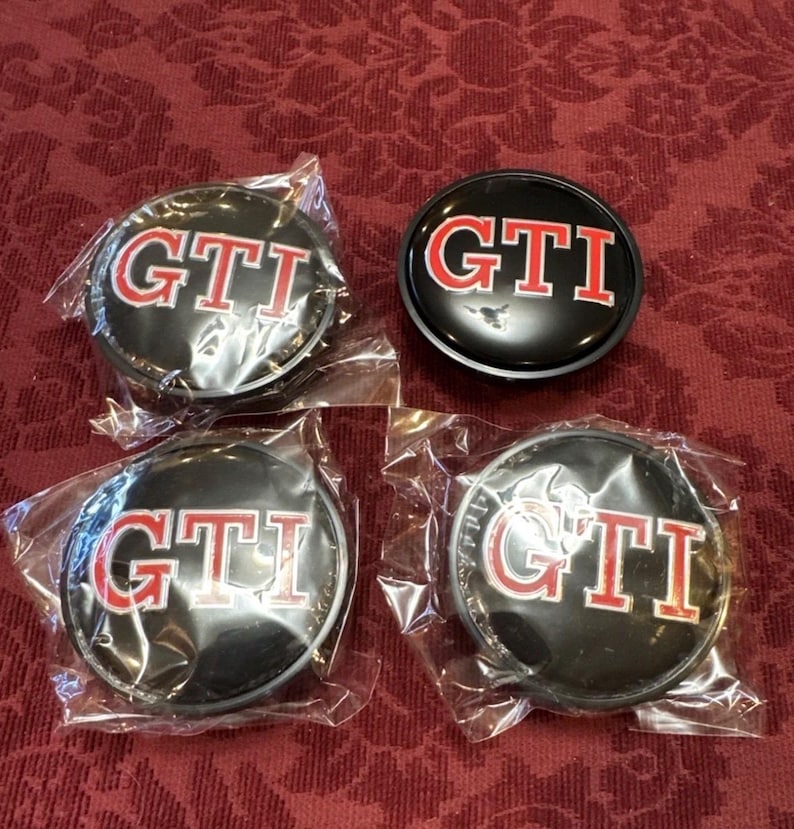 Puede incluir: Cuatro tapacubos GTI negros con letras y ribetes rojos, algunos a&uacute;n en embalaje pl&aacute;stico transparente. Las tapas son redondas y parecen ser de metal. El fondo es una tela estampada de color rojo oscuro.