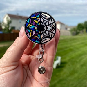 May include: A clear plastic badge holder with a round, colorful, stained glass-style design featuring a black spiderweb. The design includes shades of blue, yellow, purple, and white.