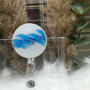 May include: A white and blue patterned badge holder with a clear retractable cord. The badge holder has a glittery finish and features a blue and white abstract design.