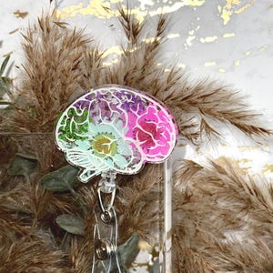 May include: A brain-shaped badge reel featuring a clear acrylic casing filled with embedded dried flowers in vibrant shades of green, light blue, and pink. The anatomical brain design is outlined in white, attached to a retractable plastic clip.