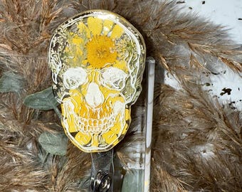 May include: A white skull with a yellow flower design encased in clear resin. The skull is attached to a clear plastic badge holder with a metal clip.