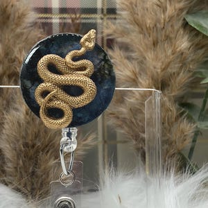 May include: A gold snake charm on a blue and black resin background. The charm is attached to a clear plastic badge holder.