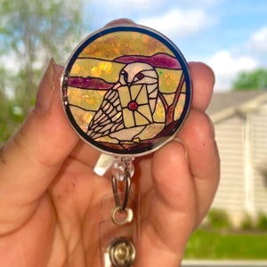 May include: A white owl with a red gem on its chest holds a letter in its talons. The owl is surrounded by a stained glass-like design with a yellow, orange, and purple background. The owl is attached to a clear plastic retractable badge holder.