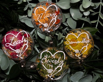 May include: Four round, clear resin badge holders with a stethoscope and heart design. Each holder features a different pressed flower design and a name: Mary, Leah, Samantha, and Beverly.