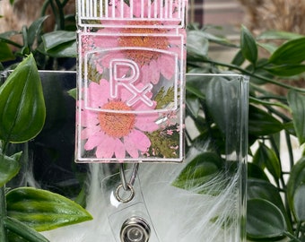 May include: A pink resin badge holder with a clear plastic backing. The front of the badge holder features a clear resin design of a prescription bottle with a pink flower inside. The design also includes a white outline of the bottle and the letter "R" with a cross.