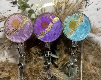 Puede incluir: Tres portagajes redondos de resina transparente con un diseño de hoja de oro y flores prensadas. Las flores son moradas, rosas y azul claro. Los portagajes están unidos a carretes de identificación retráctiles.