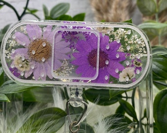 May include: A clear plastic badge holder with a purple and white pressed flower design. The holder has a metal clip on the bottom.