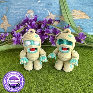 May include: Two mummy-shaped keychains with teal accents and open mouths. The keychains are beige with teal eyes, hands, and feet. They have a silver keyring attached to the top. The background includes purple flowers and green grass.