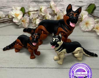 May include: Three German Shepherd dog figurines in black, brown, and cream colors. The dogs have their tongues out and are posed on a white wooden surface with white flowers in the background. The figurines are from Printverse.